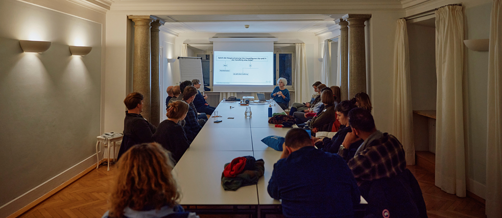 Les participant·e·s à l'atelier sont assis·es autour d'une grande table de conférence. Letícia Milano se trouve à la tête de la table et s'adresse aux participant·e·s.  