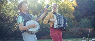 ein Mann spielt auf einer Art Gitarre und singt, neben ihm eine Frau, die Akkordeon spielt und mitsingt, die beiden stehen in der Natur 