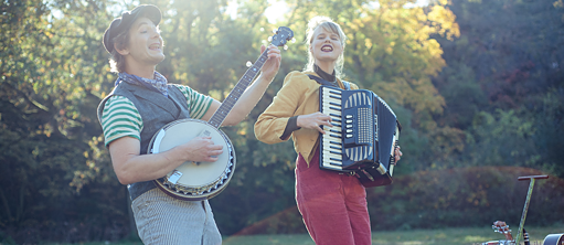 ein Mann spielt auf einer Art Gitarre und singt, neben ihm eine Frau, die Akkordeon spielt und mitsingt, die beiden stehen in der Natur 