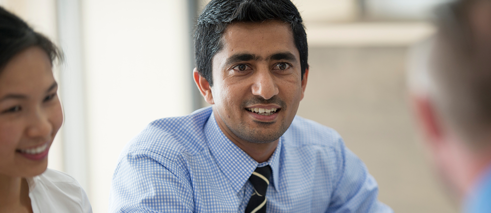 A person in a tie and checkered shirt smiles during a discussion, with others partially visible in the background.