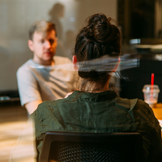 Two people sitting across from each other at a table, one with their back to the camera and the other out of focus in the background.