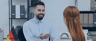  A smiling person in a light blue shirt gestures while speaking to another person with reddish hair in an office setting. © © Goethe-Institut Housing in Germany