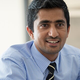 A person in a tie and checkered shirt smiles during a discussion, with others partially visible in the background.