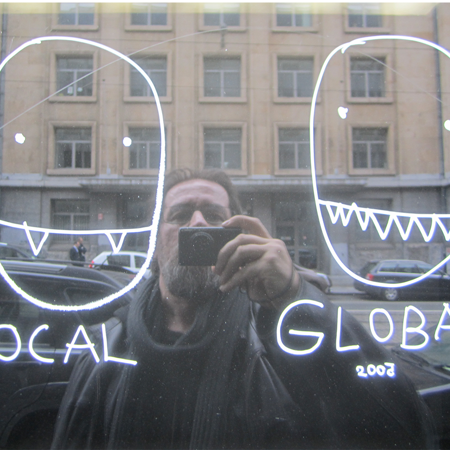  Dan Perjovschi macht in einem spiegelnden Fenster ein Selfie; auf dem Fenster sind links und rechts Köpfe skizziert mit den Unterschriften local und global